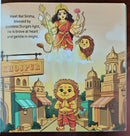 A children's book or printed card featuring an illustrated scene with a crowned goddess in red and gold standing on clouds above a lion, with a smaller lion child in front of a city street labeled Khajur. Text on the left reads, 'Meet Bal Simha, blessed by Goddess Durga's light, He is brave at heart and gentle in might.
